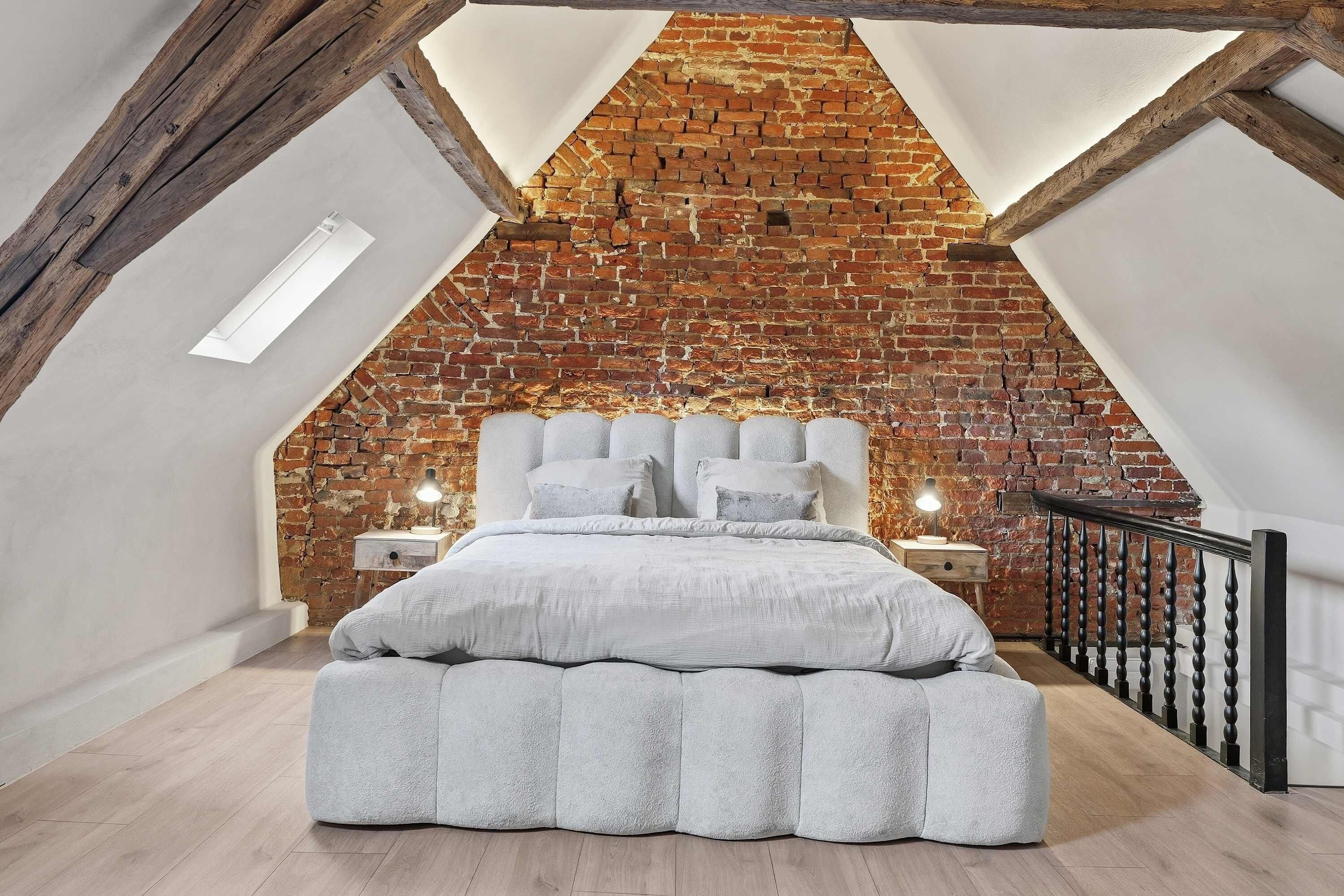 A loft bedroom with exposed brick walls, a sloped ceiling, and a large bed with a fluffy white comforter.