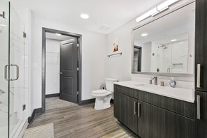 A modern bathroom with a glass shower enclosure, a white toilet, and dark wood cabinets. - NV Hotel w/ Pool, Gym, Rooftop Lounge & Free Park (Nashville)