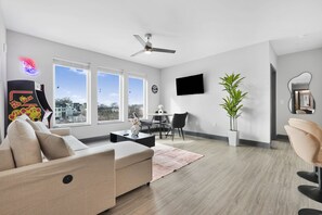 A modern living room with a sofa, coffee table, and a dining area with chairs. There is a TV mounted on the wall, a ceiling fan, and large windows offering a view of the outside. - NV Hotel w/ Pool, Gym, Rooftop Lounge & Free Park (Nashville)