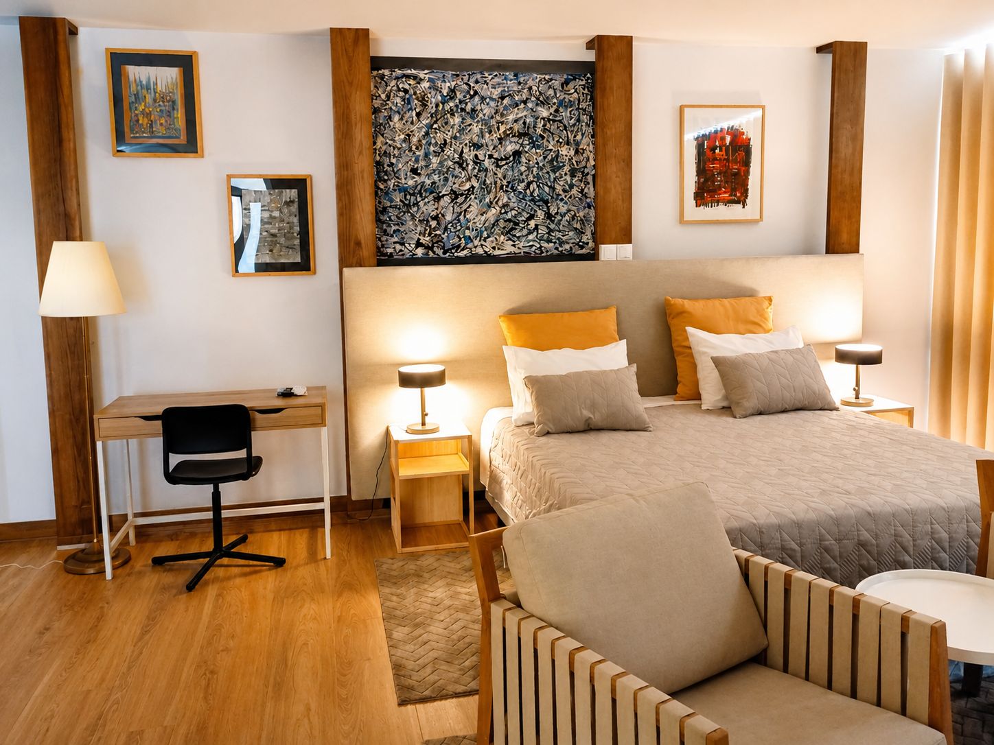 A hotel room with a bed, bedside tables, a desk, a chair, and artwork on the walls.