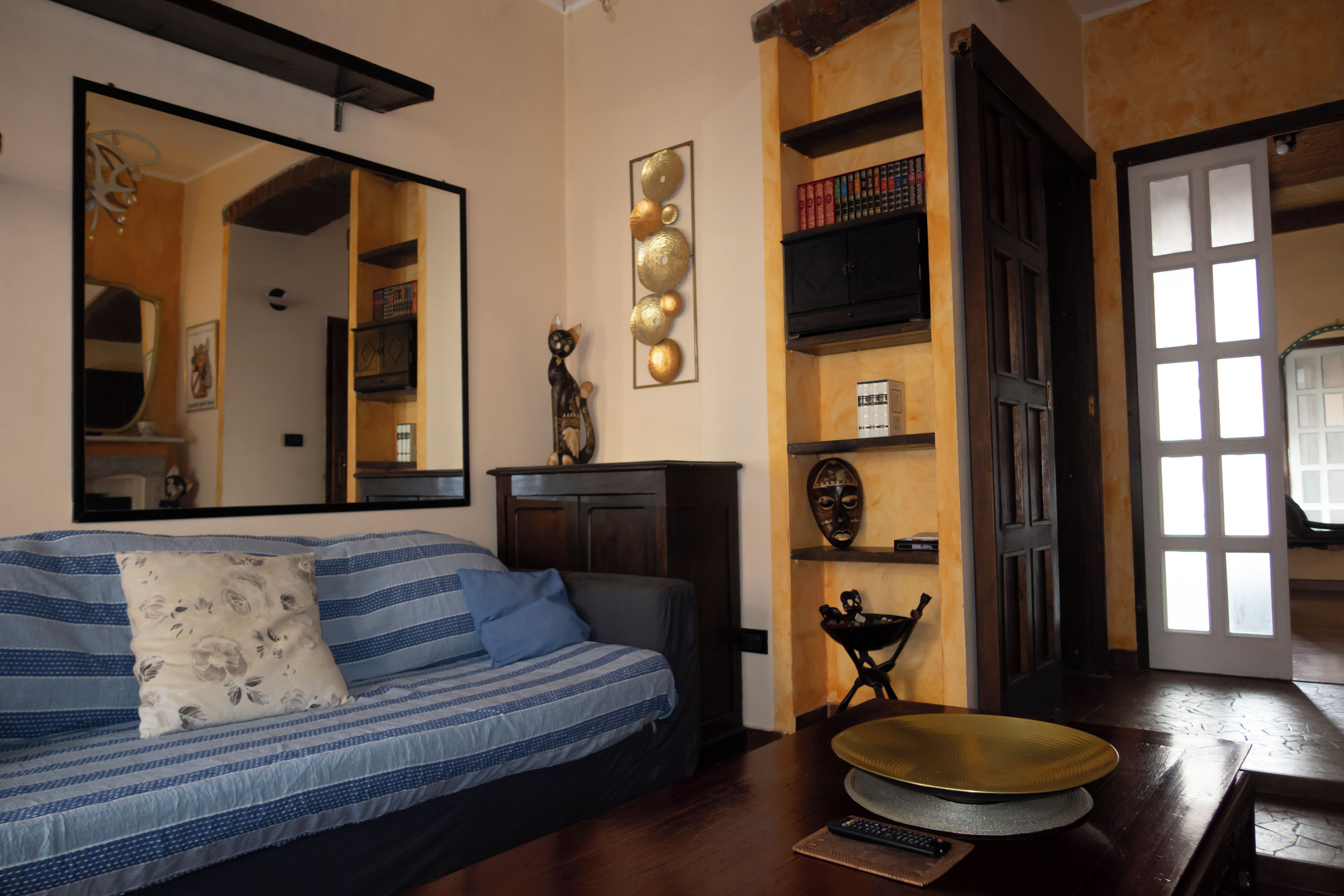 Una habitación con sofá, espejo, estante con libros, una máscara decorativa y una puerta con ventanas.