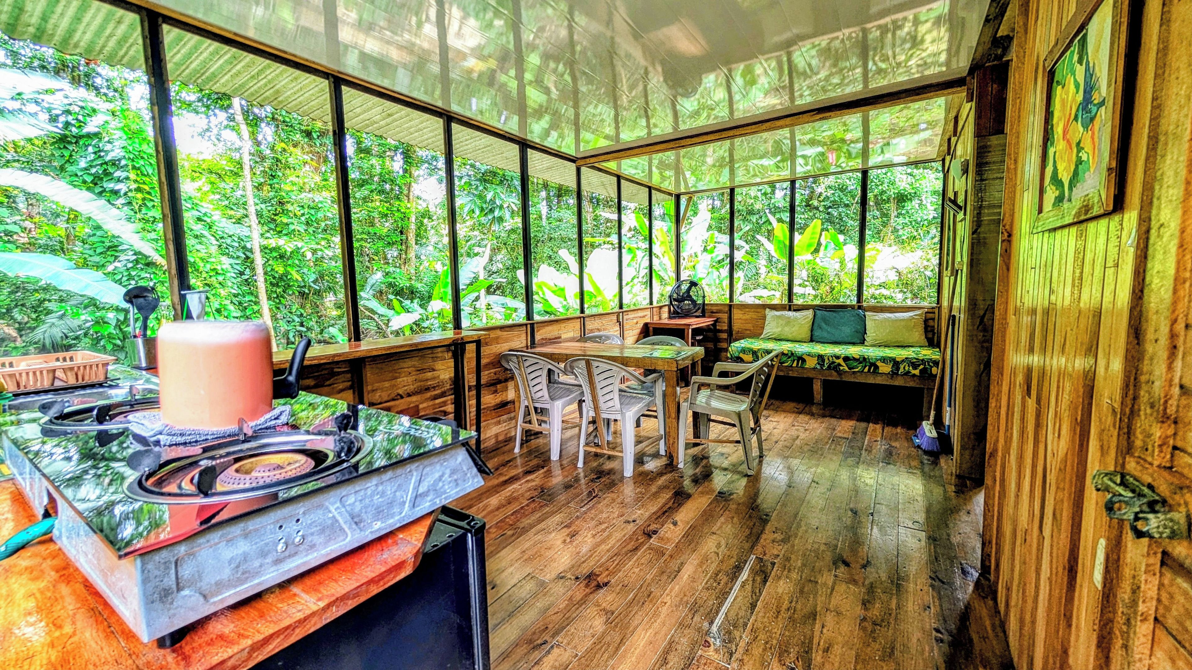 Una habitación con piso de madera, una mesa de comedor con sillas, un sofá y un ventanal con vista a la vegetación.