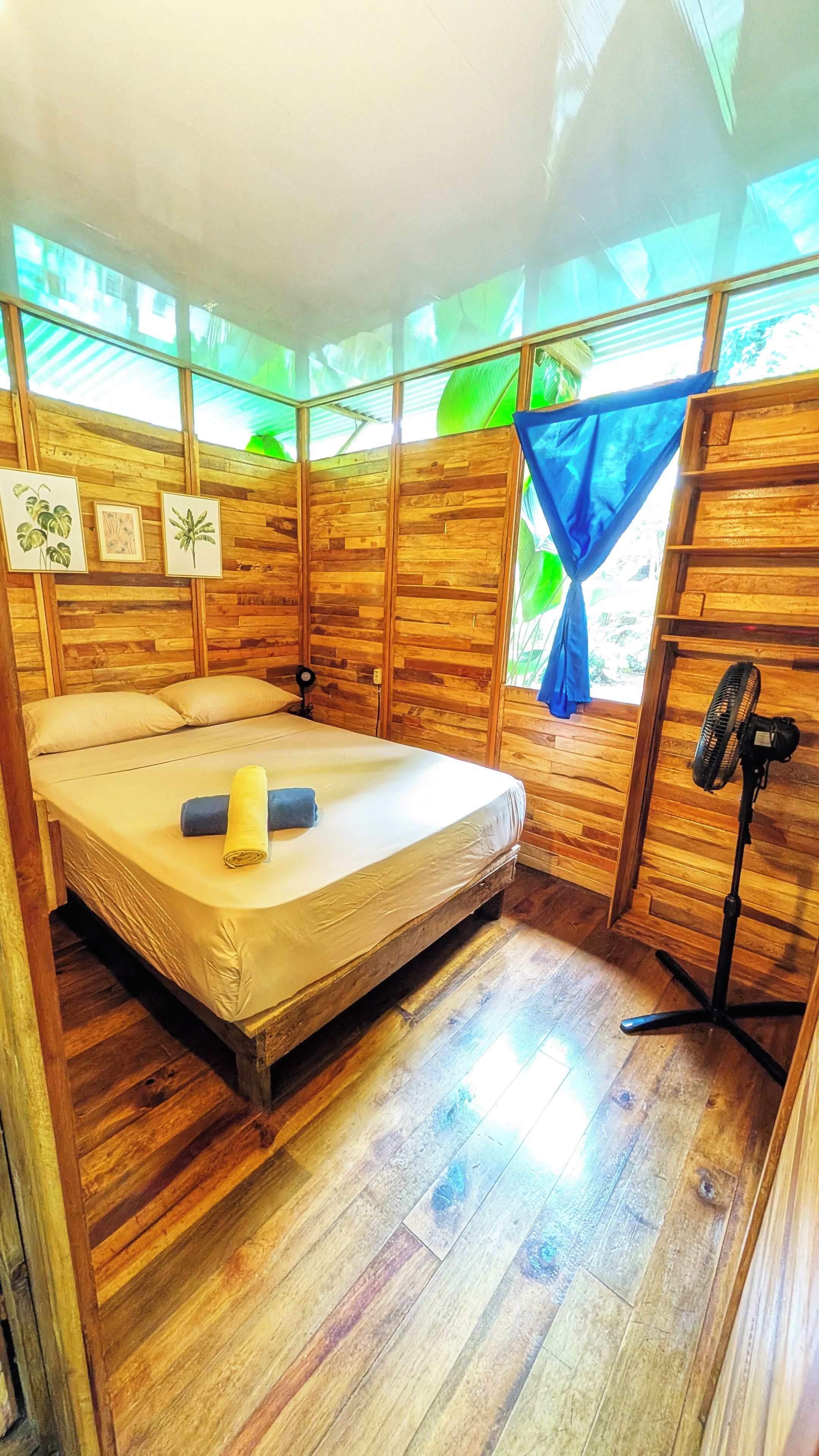 Una habitación con una cama, un ventilador de pie y paredes de madera.