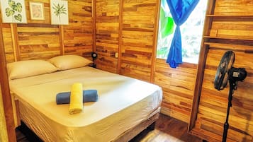 Una habitación con una cama, un ventilador de pie y paredes de madera.
