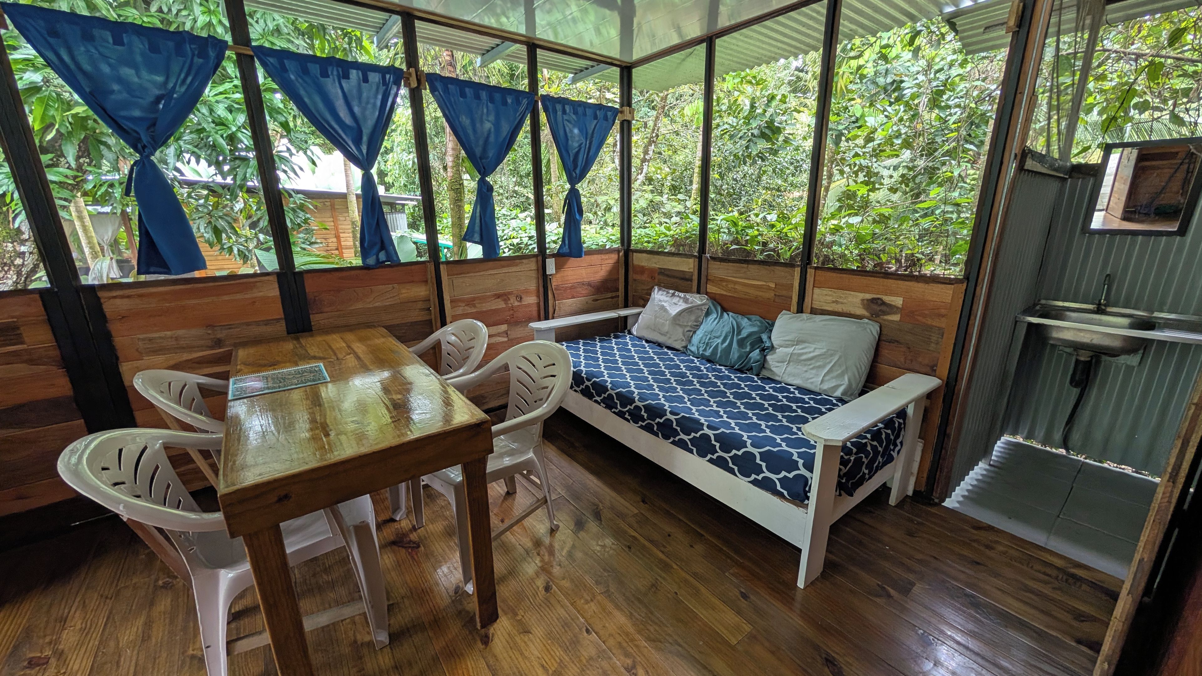 Una habitación con piso de madera, cama con ropa de cama estampada, una mesa con sillas y una ventana con cortinas.