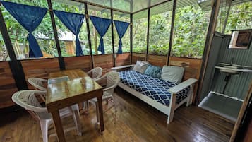 Una habitación con piso de madera, cama con ropa de cama estampada, una mesa con sillas y una ventana con cortinas.