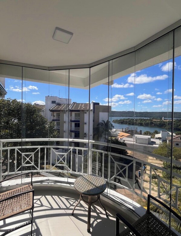 A balcony with a glass railing, a round table, and chairs, overlooking a body of water and buildings. - New Listing (Caldas Novas)