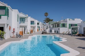 A swimming pool area with a clear blue pool, white buildings with green accents, and a palm tree in the background. - 149 San Fermin, A/C, 500Mbps, Parking, 9 Mins to Beach, Ground Floor (Puerto del Carmen)