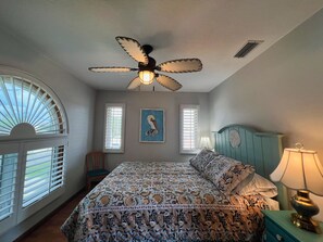 Ein Schlafzimmer mit einem Bett, einem Deckenventilator, einem Fenster mit Rollläden, einem Wandbild und einem Nachttisch mit Lampe.