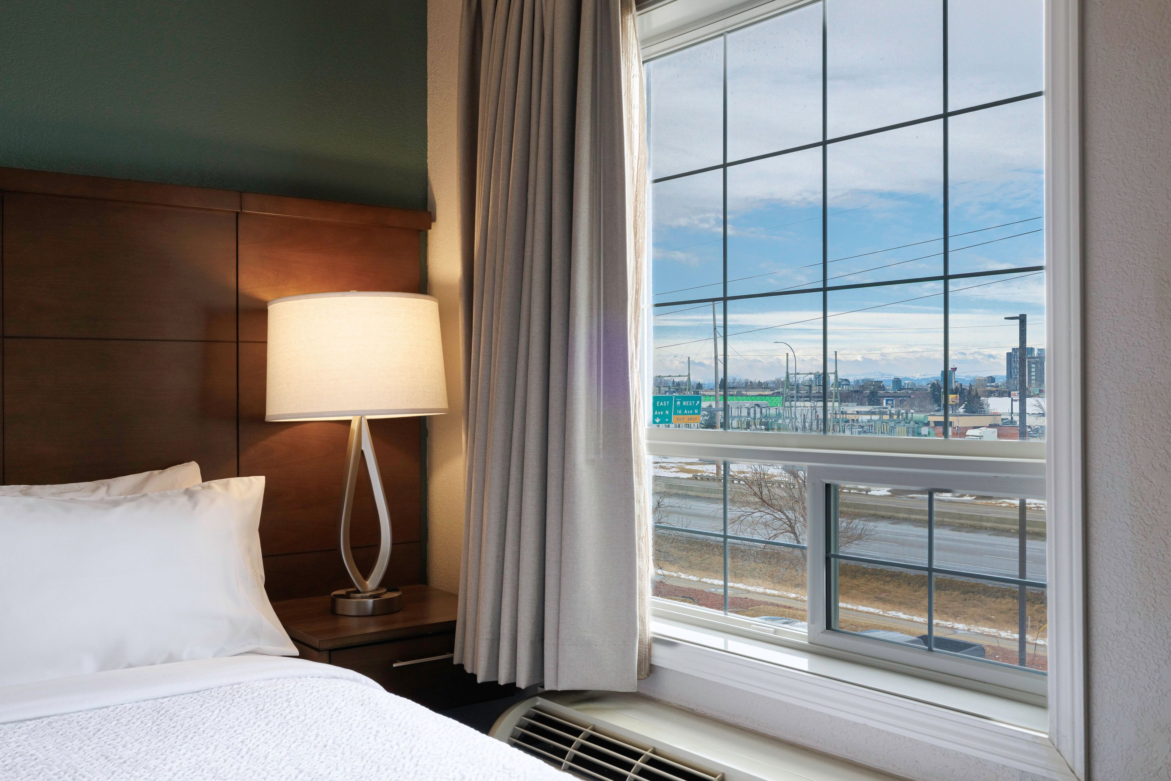 A hotel room with a bed, a bedside lamp, a window with a view of a cityscape.