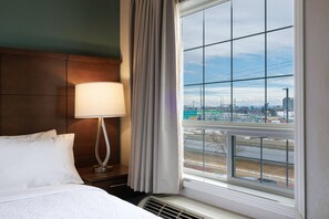 Habitación de hotel con cama, lámpara de noche y ventana con vistas a un paisaje urbano.