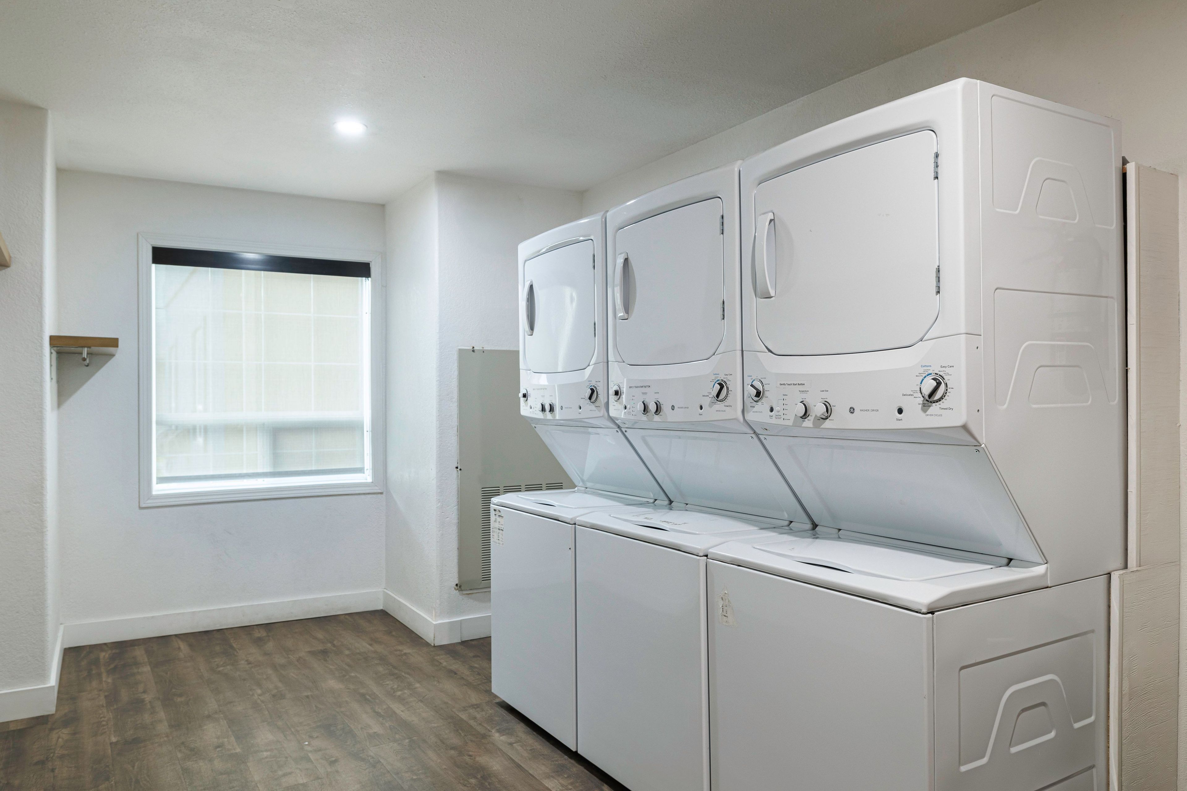 A laundry room with three stacked washers and dryers, a window with blinds, and a wooden floor.