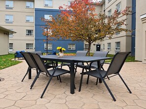 Un patio exterior con una mesa y sillas, un árbol con hojas otoñales y un edificio al fondo.