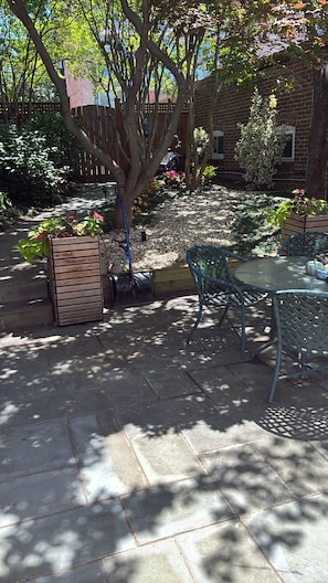 Un patio con mesa y sillas, rodeado por un jardín con diversas plantas y un árbol.