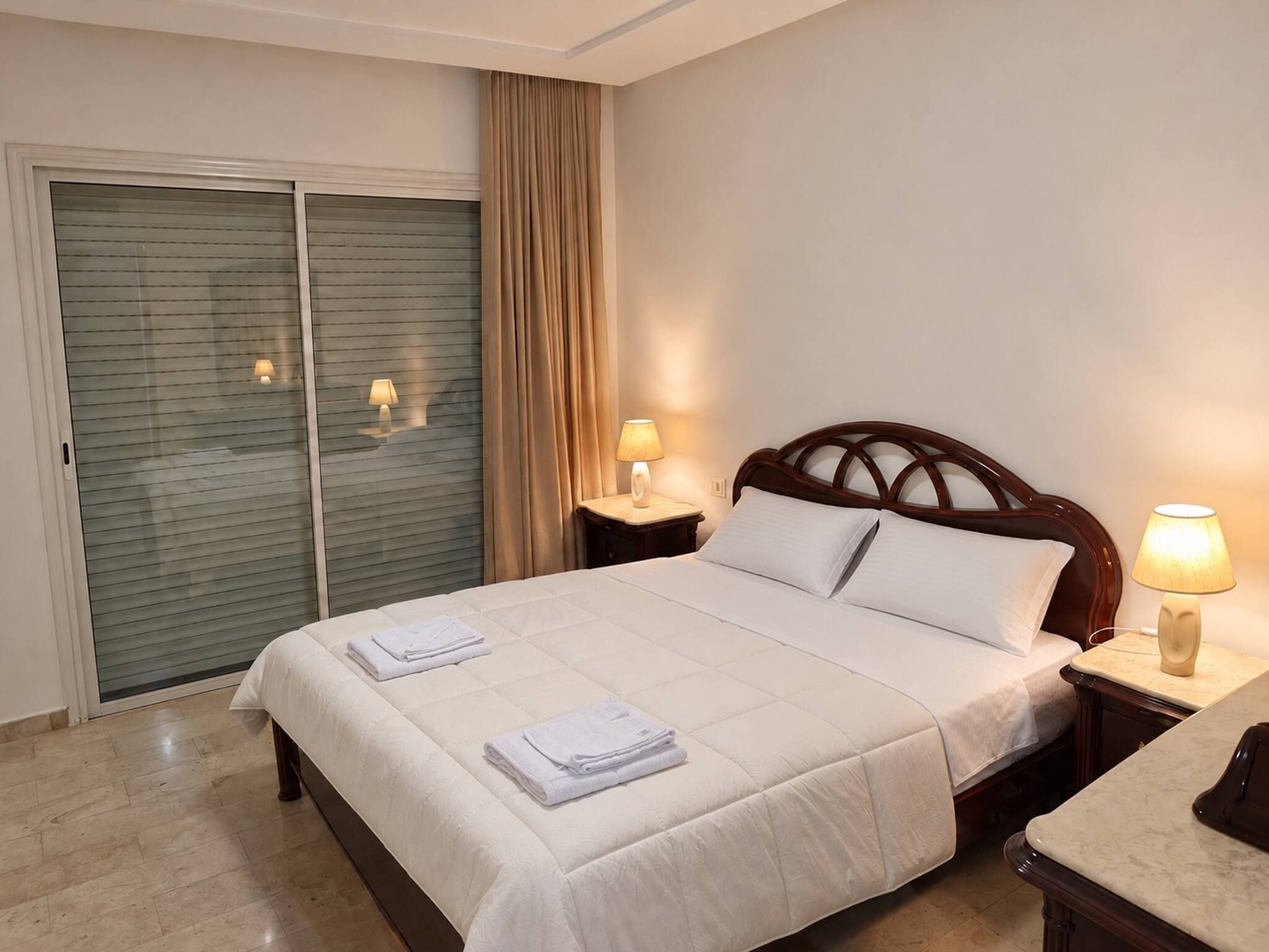 A hotel room with a bed, two bedside lamps, a wooden headboard, a large window with curtains, and a sliding door.