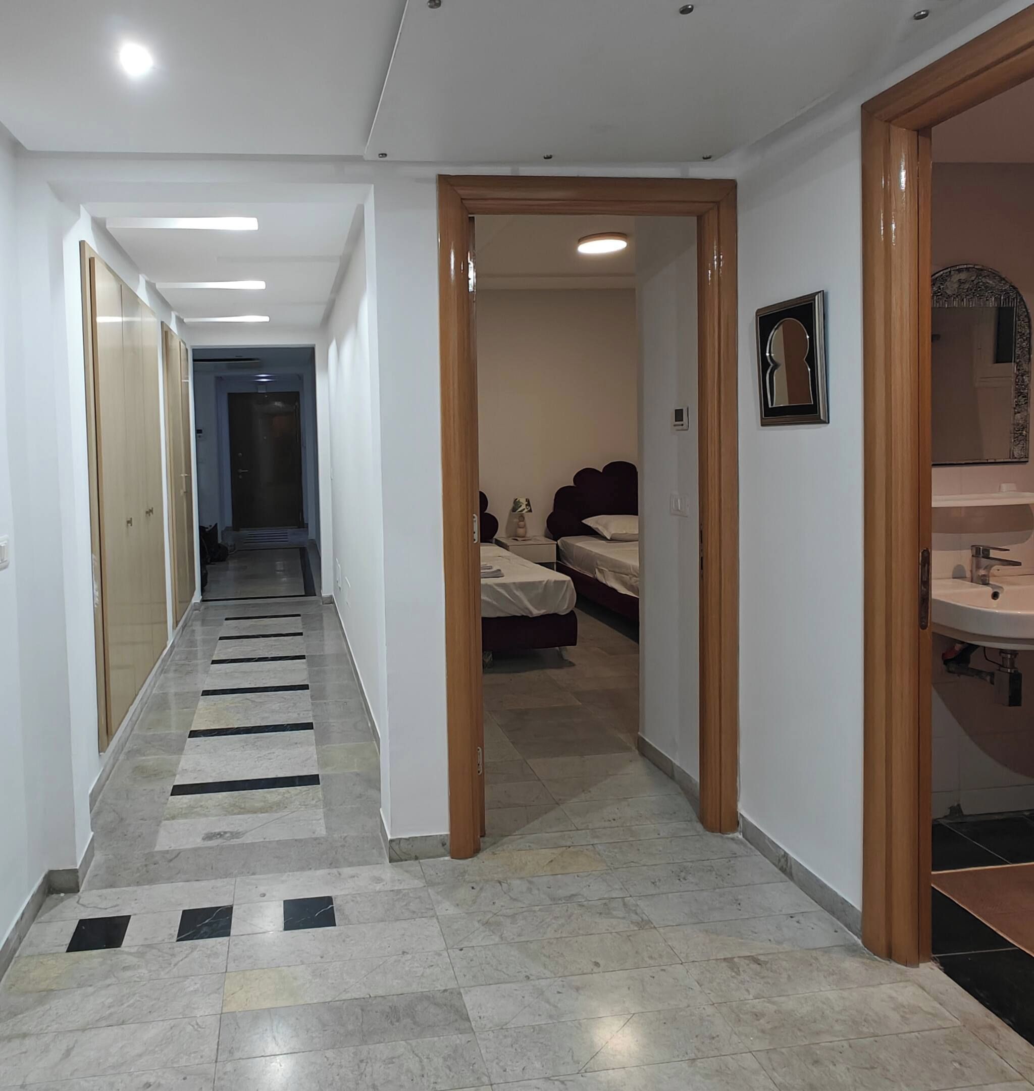 A hallway with a tiled floor leading to a bedroom with a bed and a bathroom with a sink.