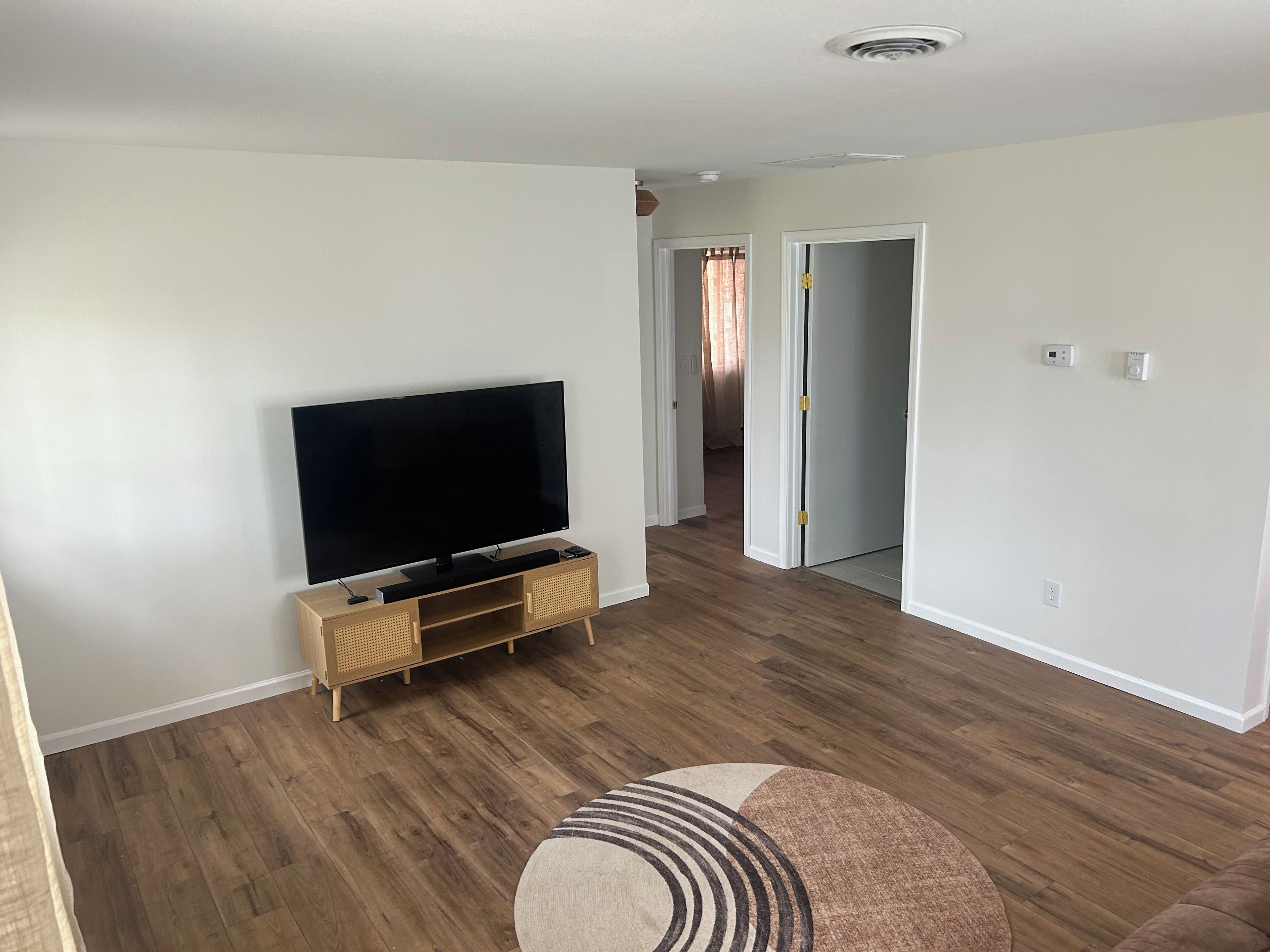 Ein Wohnzimmer mit einem Flachbildfernseher auf einem Holzboden, einem runden Teppich und einem Flur, der in ein weiteres Zimmer führt.
