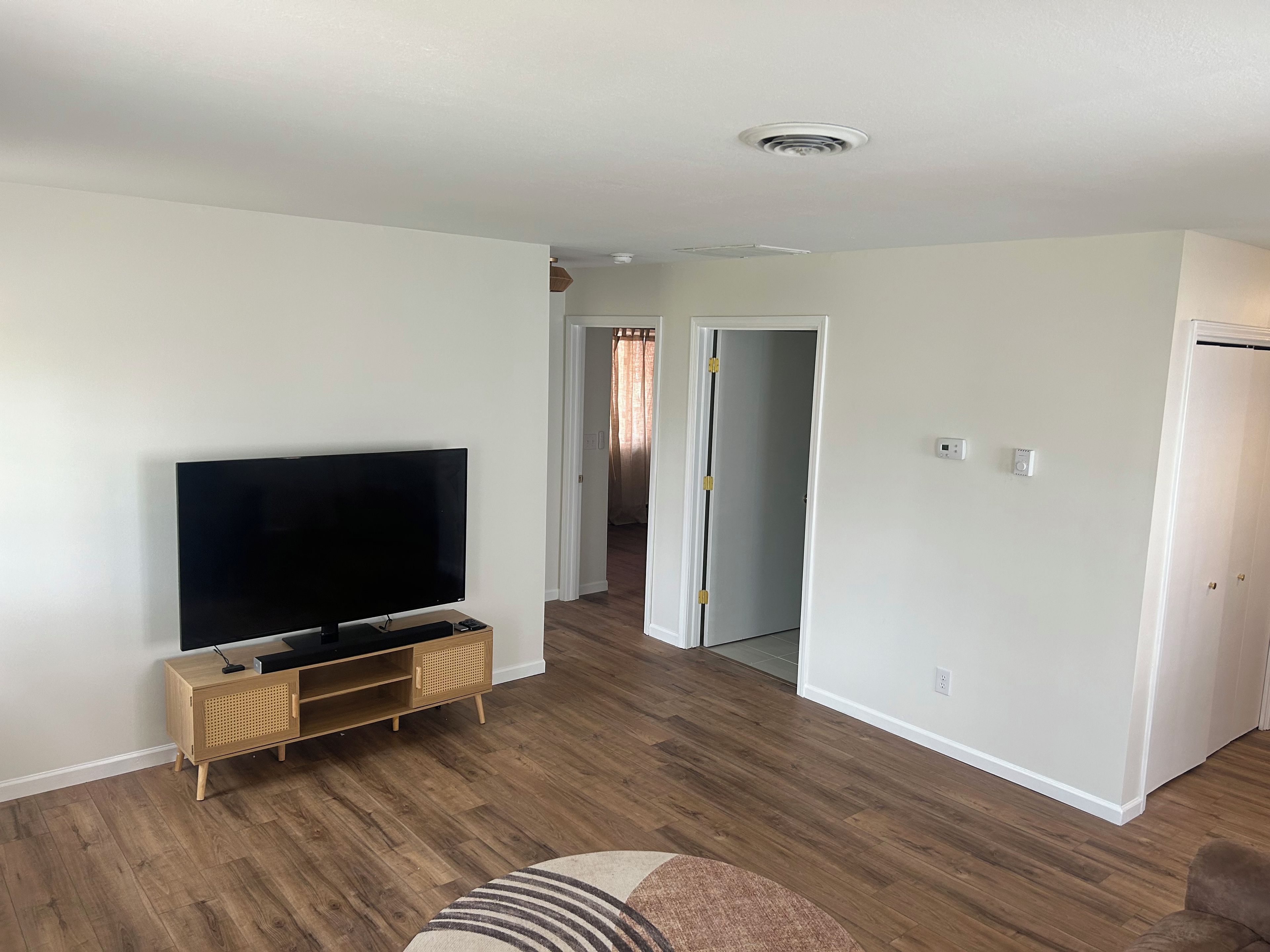 Una sala de estar con una televisión de pantalla plana sobre un soporte de madera, un pasillo que lleva a otra habitación y una puerta con manija.