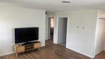 Una sala de estar con una televisión de pantalla plana sobre un soporte de madera, un pasillo que lleva a otra habitación y una puerta con manija.
