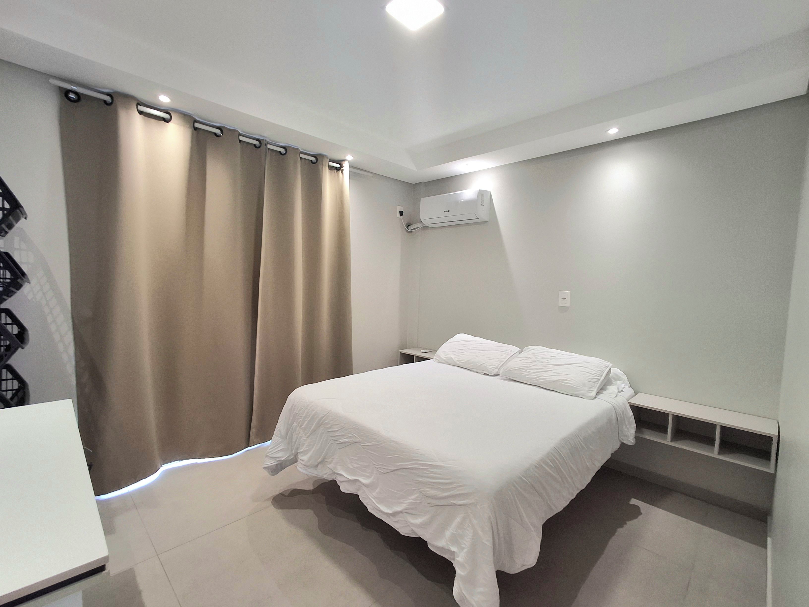 Un dormitorio con cama, cortinas, aire acondicionado y una mesita de noche blanca.