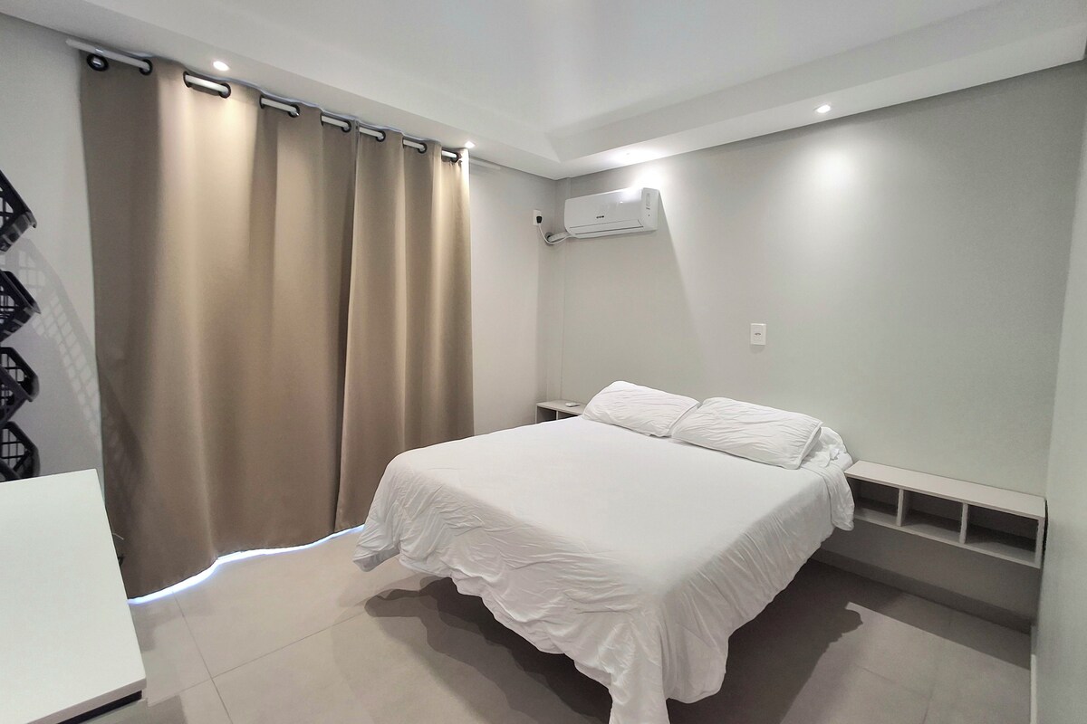 Un dormitorio con cama, cortinas, aire acondicionado y una mesita de noche blanca.