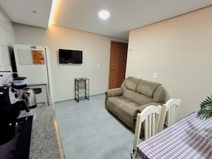 Un salón con un sofá, una mesita, un televisor montado en la pared y una zona de cocina con hervidor y cafetera.