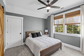 Une chambre à coucher comprenant un lit, un ventilateur de plafond, une table de chevet, une fenêtre avec des stores et une porte.