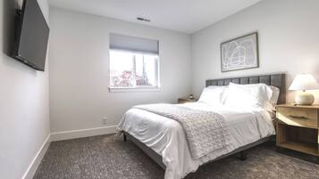 A bedroom with a bed, a bedside table with a lamp, a window with blinds, and a wall-mounted TV.