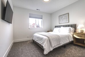 A bedroom with a bed, a bedside table with a lamp, a window with blinds, and a wall-mounted TV. - Bright Modern Home, Sauna, King, 1.9 mi. to Autzen (Eugene)