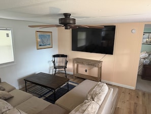 Een woonkamer met een plafondventilator, een aan de muur bevestigde televisie, een houten salontafel, een beige bank en een fauteuil.