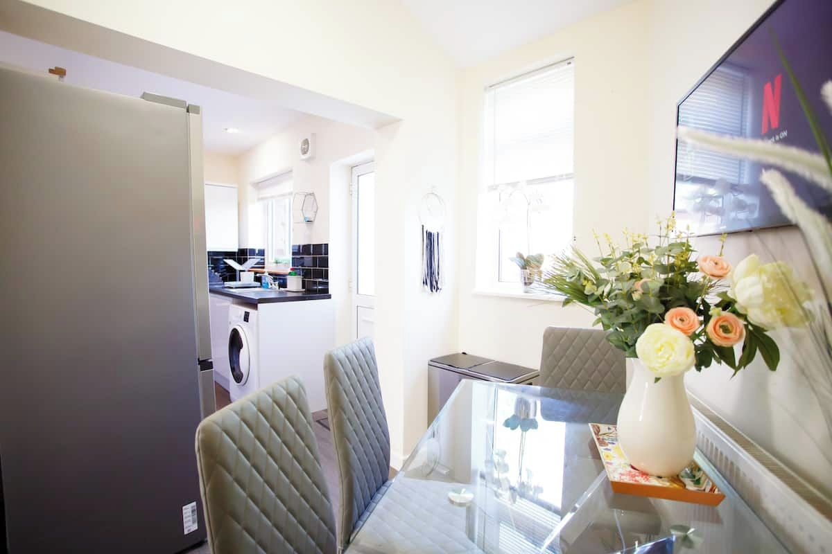A dining area with a glass table, cushioned chairs, a vase with flowers, and a kitchen in the background.