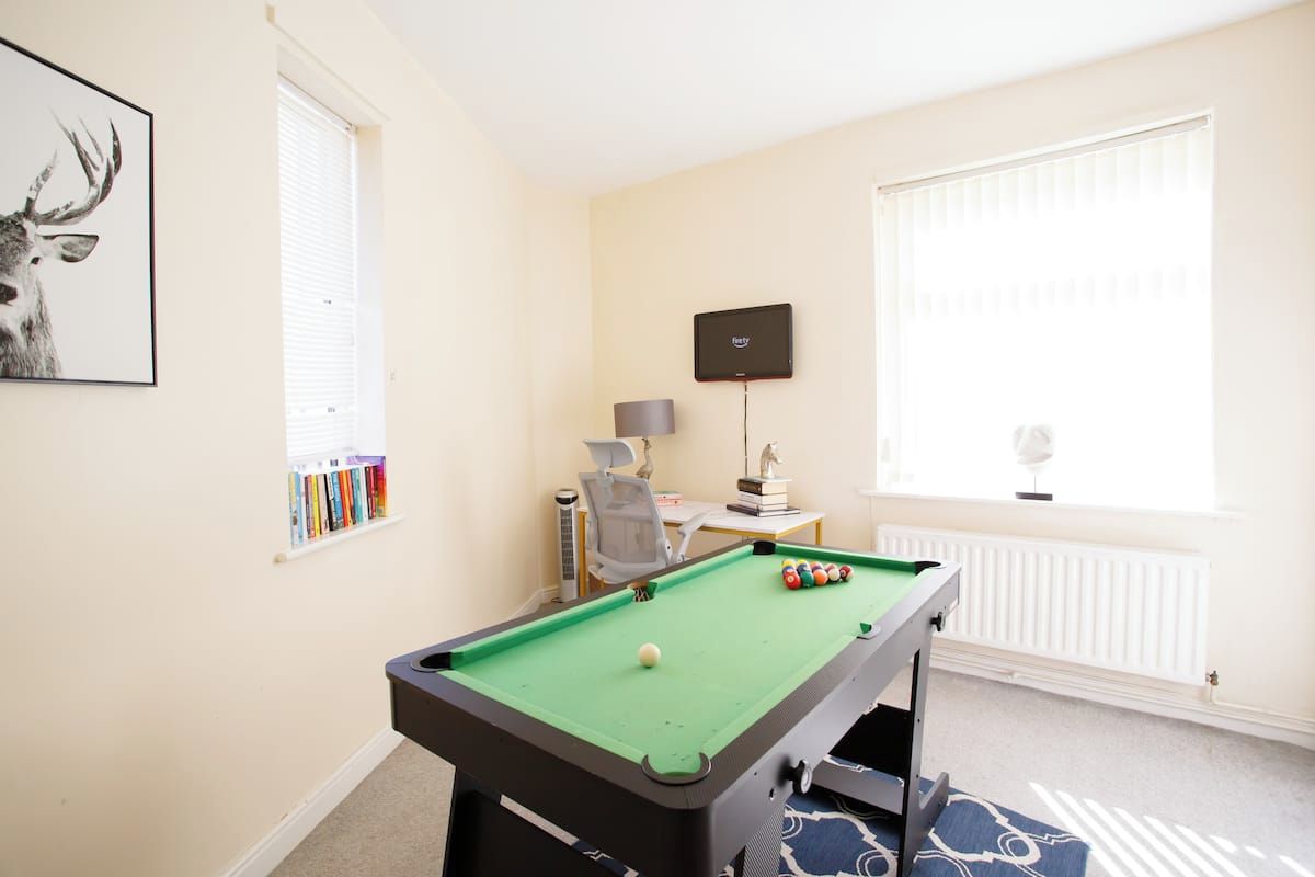 Une pièce comprenant une table de billard, une bibliothèque, un bureau avec un ordinateur et une fenêtre avec des stores.