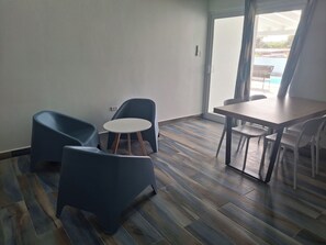 Een kamer met een eettafel en stoelen, een kleine ronde tafel met stoelen, en uitzicht door een glazen deur op een buitengebied.