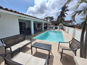 Un espace piscine avec des chaises longues et une table, entouré d’une terrasse couverte et de palmiers.