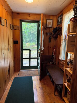 Interior de una cabaña de madera con un banco, una estantería con objetos y una puerta que da al exterior.