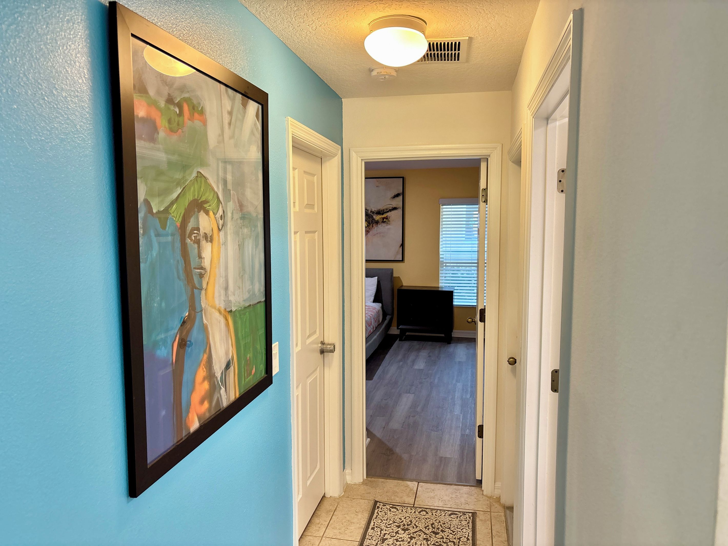 A hallway with a painting on the wall, a door leading to a bedroom, and a ceiling light.
