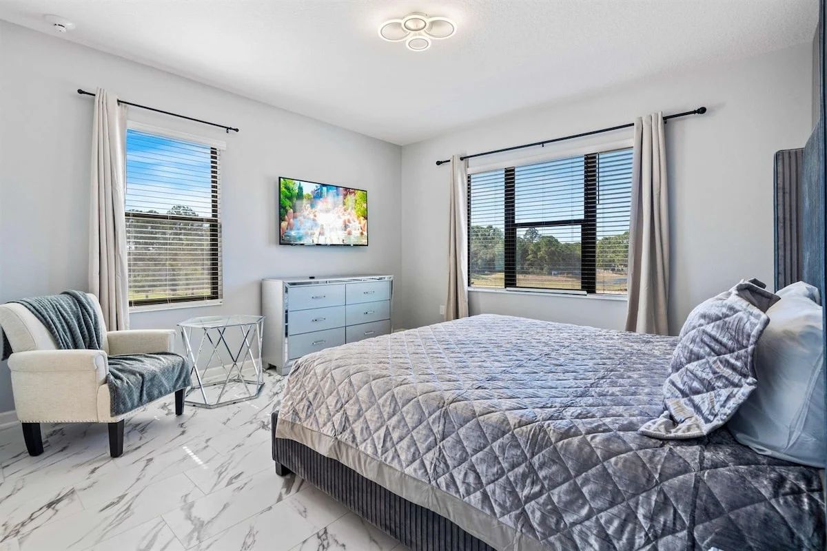A bedroom with a large bed, a chair, a small table, and a TV on the wall.
