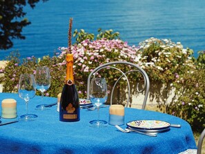 Et bord dækket til to med vin, glas og tallerkener, foran en have med blomster og havudsigt.