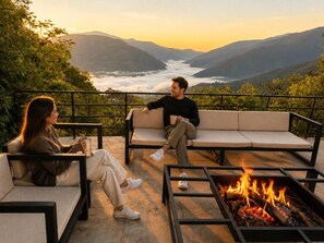 A couple seated on a sofa with a fire pit in front, overlooking a valley.
