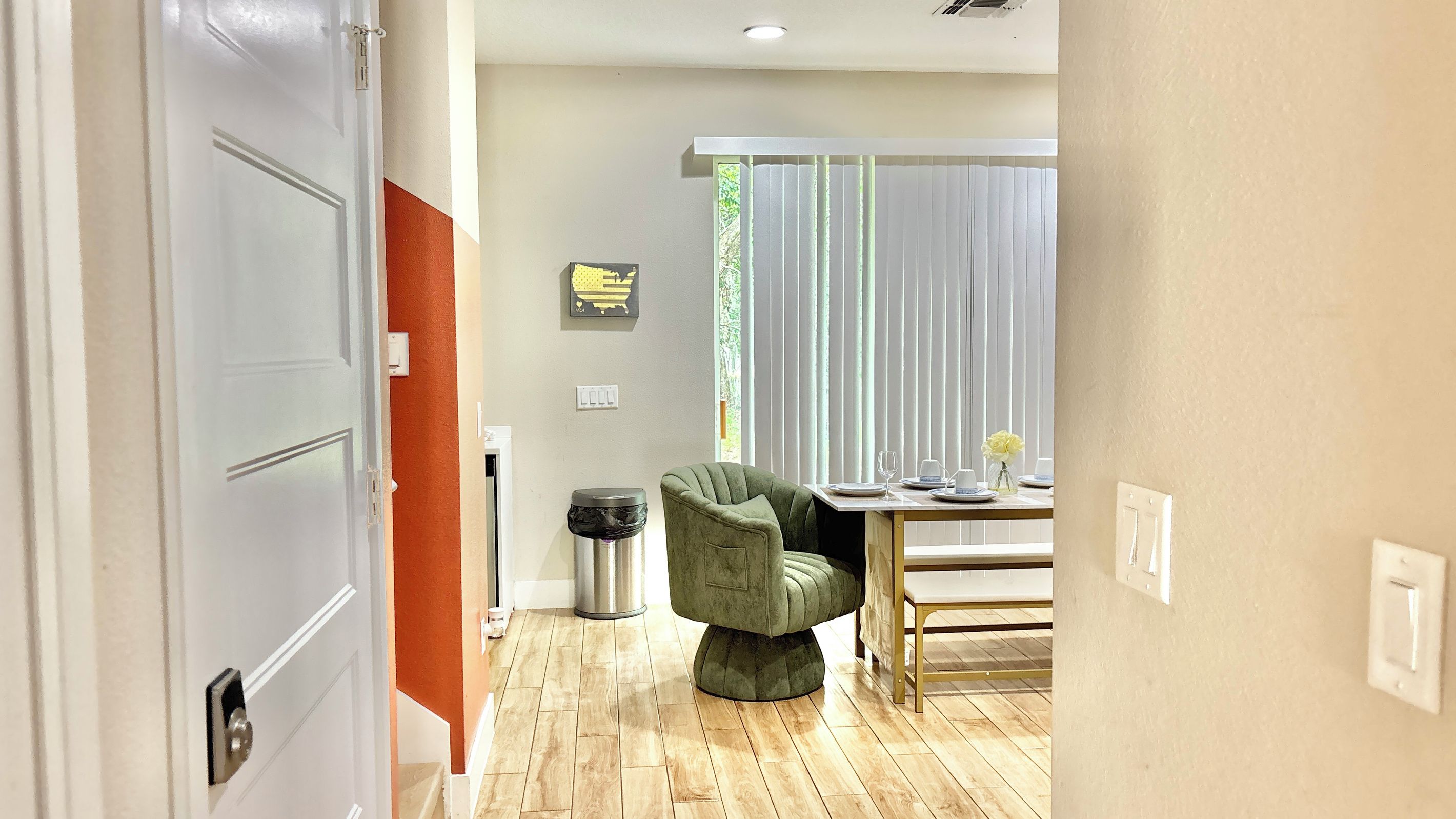 A hotel room with a dining area, a green upholstered chair, a table set with plates and glasses, and a window with blinds.