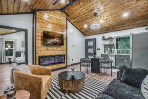Un salon moderne avec une cheminée, un plafond en bois et un coin repas donnant sur une cuisine en arrière-plan.