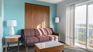 Ein modernes Hotelzimmer mit einer Couch, einem Beistelltisch, einer Lampe und Blick auf die Stadt durch große Fenster.