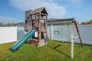 Une structure de jeu en bois, dotée d’un toboggan et de balançoires, se dressant devant une clôture blanche et sous un ciel dégagé.