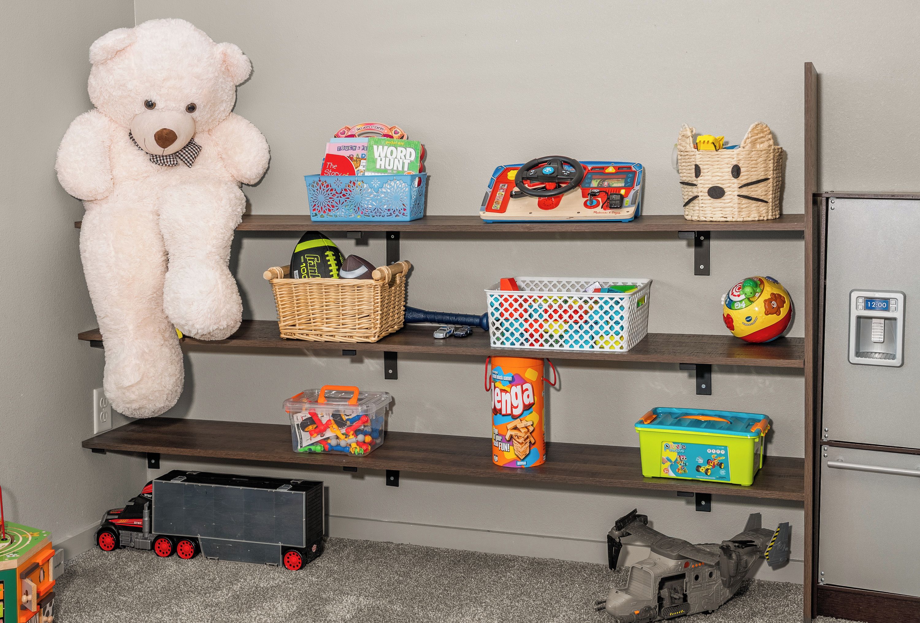 Een kamer met een groot knuffelbeer, planken vol speelgoed en spellen, een speelgoedvrachtwagen en een klein speelgoedvliegtuig.