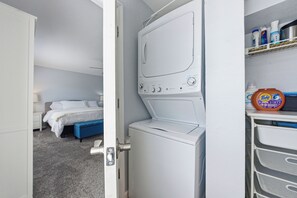 Zona de bugaderia amb rentadora, un llit i prestatges amb productes de neteja.