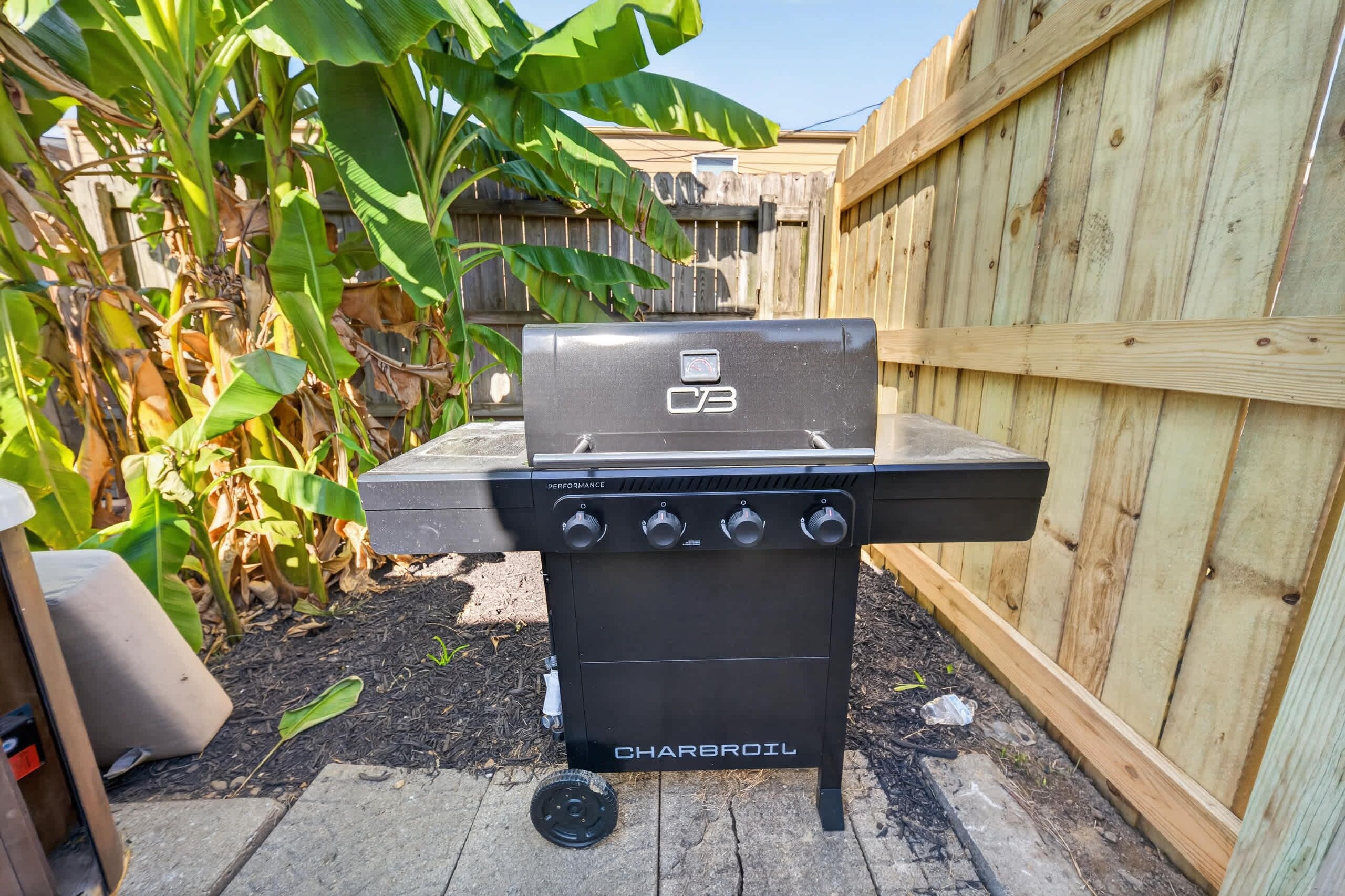 Ein Charbroil-Grill mit vier Bedienknöpfen steht auf einer Betonpatio mit Mulch, neben einem Holzzaun und einer Bananenpflanze.