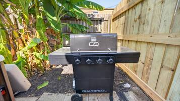 Ein Charbroil-Grill mit vier Bedienknöpfen steht auf einer Betonpatio mit Mulch, neben einem Holzzaun und einer Bananenpflanze.