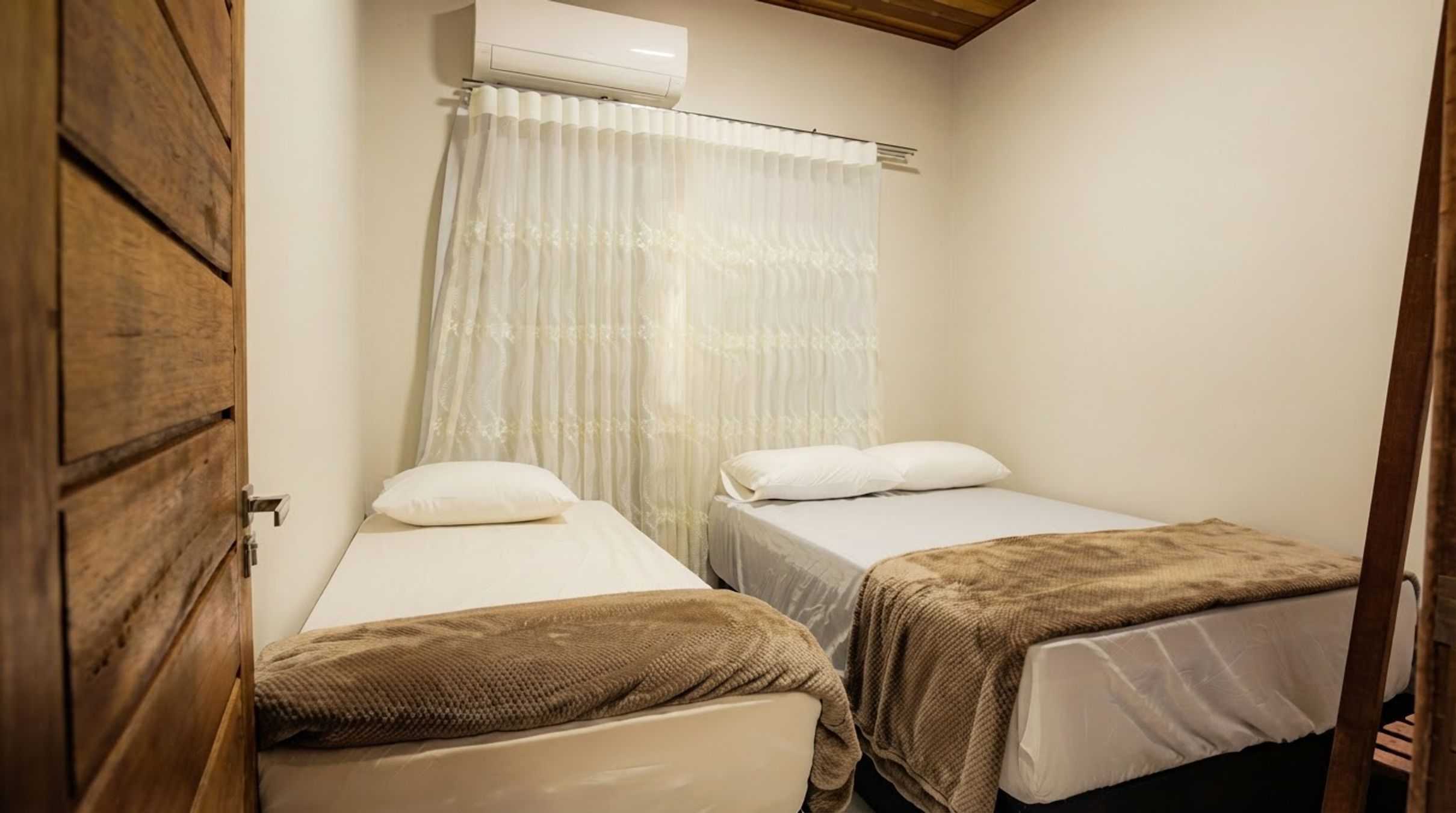 Um pequeno alojamento de um quarto, com duas camas, uma porta de madeira e uma janela com cortinas leves.
