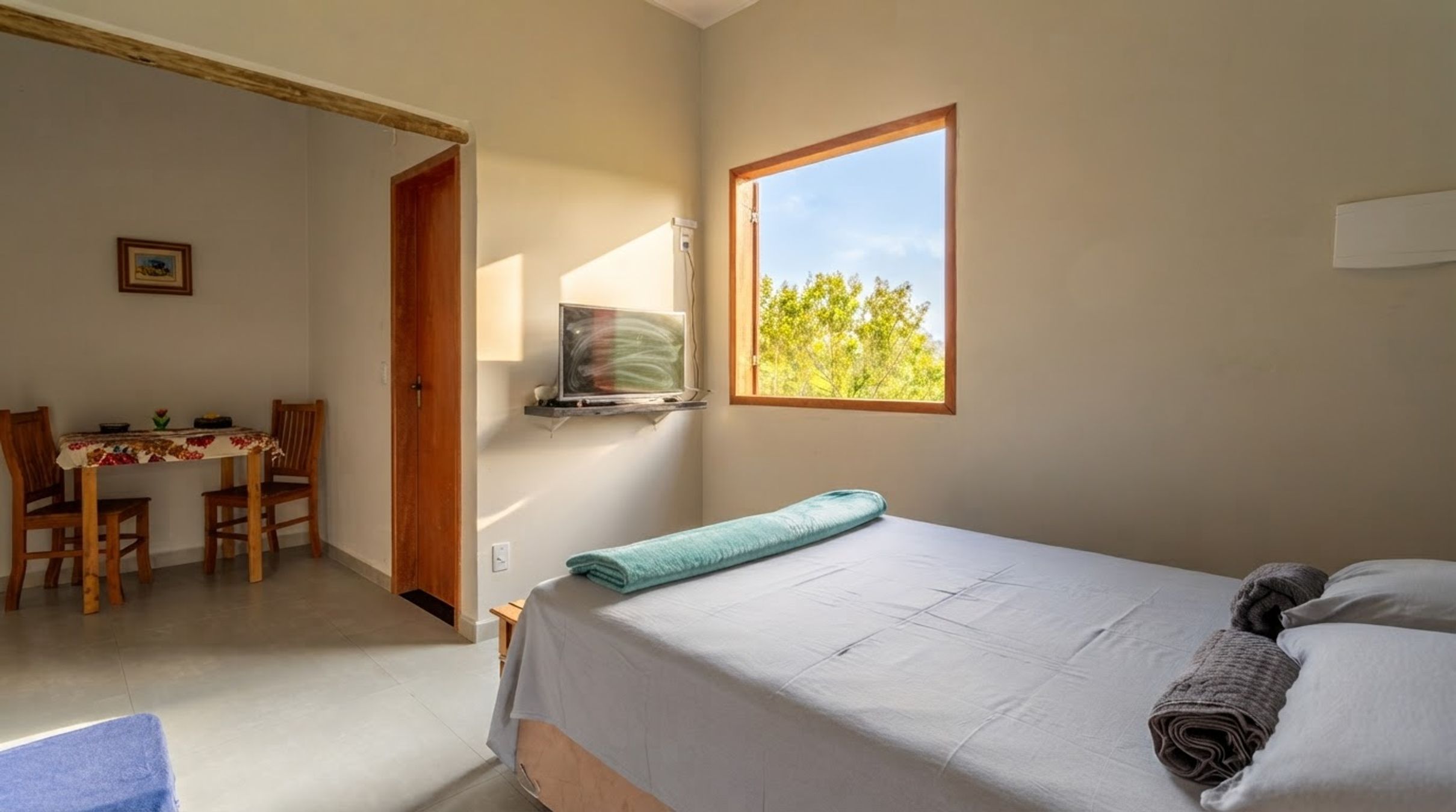 Ein Schlafzimmer mit Bett, einem Fenster mit Blick auf Bäume, einem Schreibtisch mit Stühlen und einer Tür zu einem anderen Raum.
