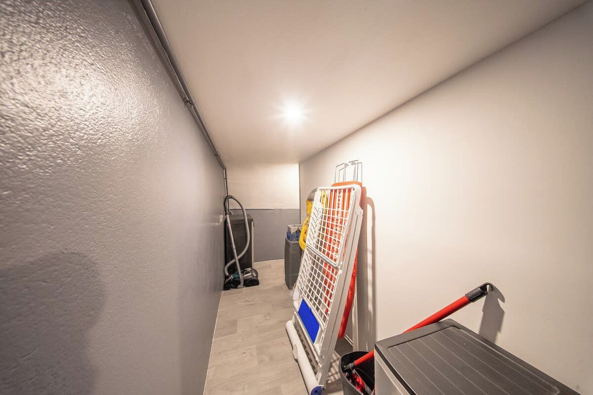 A narrow hallway with a ceiling light, a vacuum cleaner, and a red-handled tool leaning against the wall.